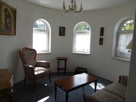 Um quarto com um lustre, duas janelas em arco, uma cadeira de madeira, uma mesa pequena e um sofá.