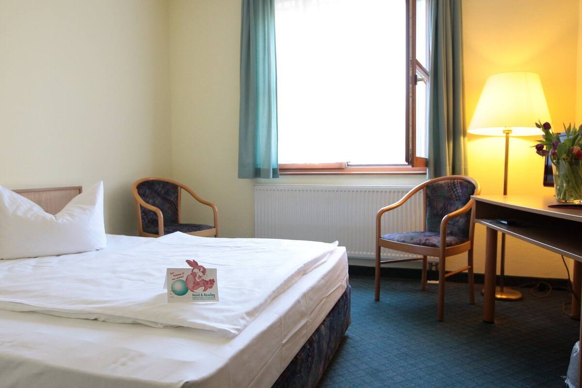A hotel room with a bed, two chairs, a desk, a lamp, and a window with curtains.
