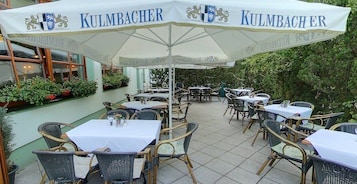 Un área de estar al aire libre con mesas y sillas bajo un paraguas grande con la marca 'KULMBACHER'.