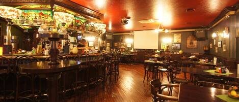 A bar area with wooden flooring, bar stools, and a variety of bottles and glasses on shelves.