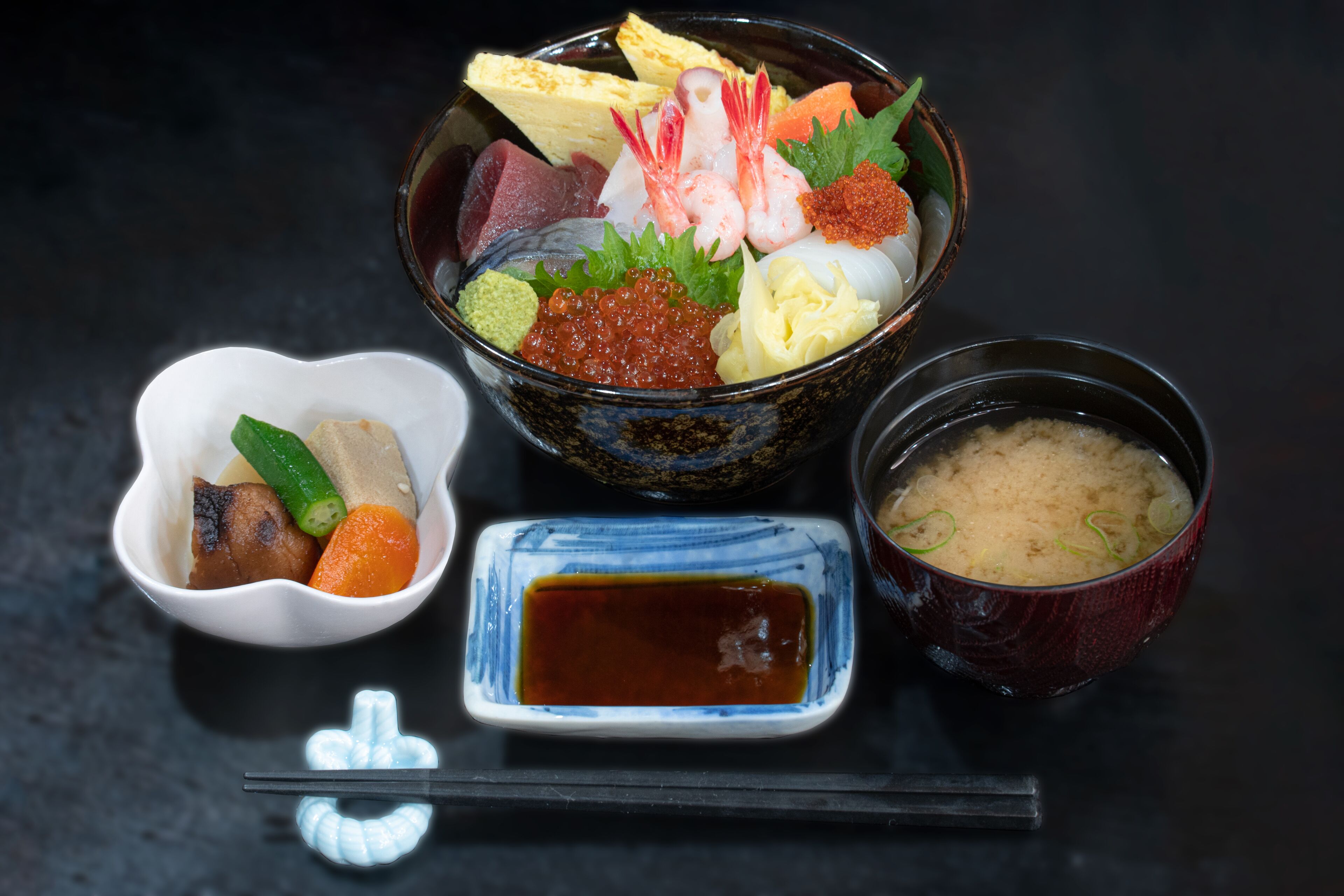 Un tazón de sashimi con diferentes ingredientes, un recipiente pequeño de vegetales encurtidos, un tazón de sopa de miso y un recipiente pequeño de salsa de soya.