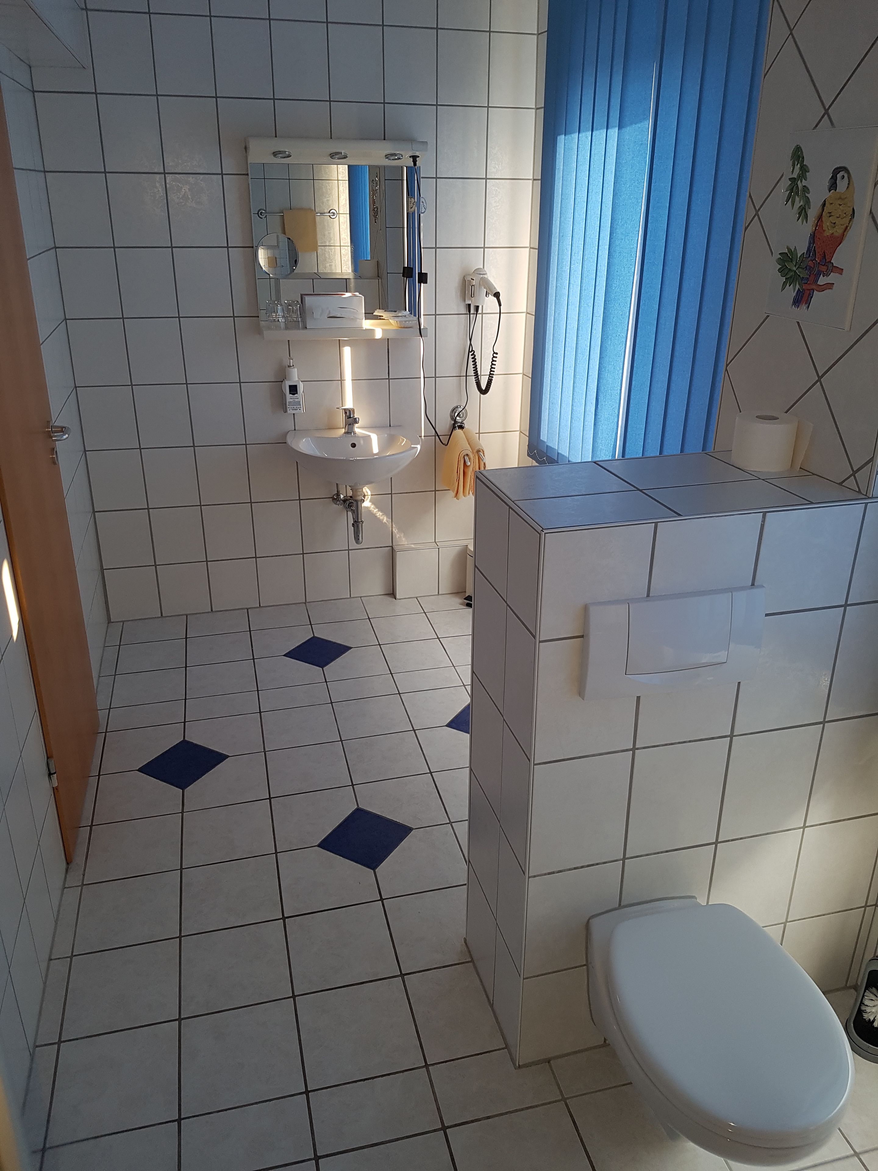 Un baño con paredes de azulejos blancos, un lavamanos blanco, un inodoro y una cortina de ducha azul.