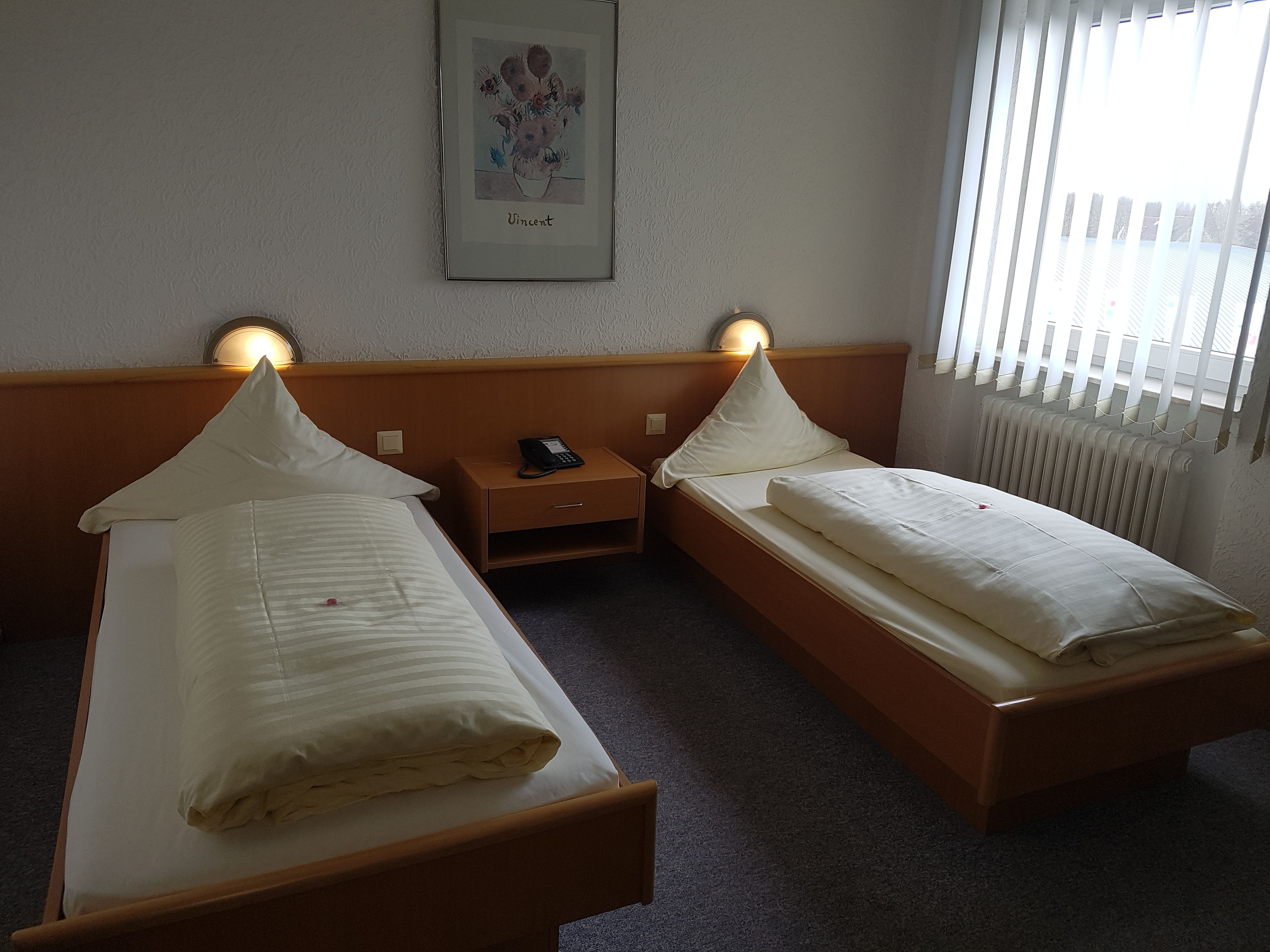 A hotel room with two single beds, a bedside table with a phone, a framed artwork on the wall, and a radiator.