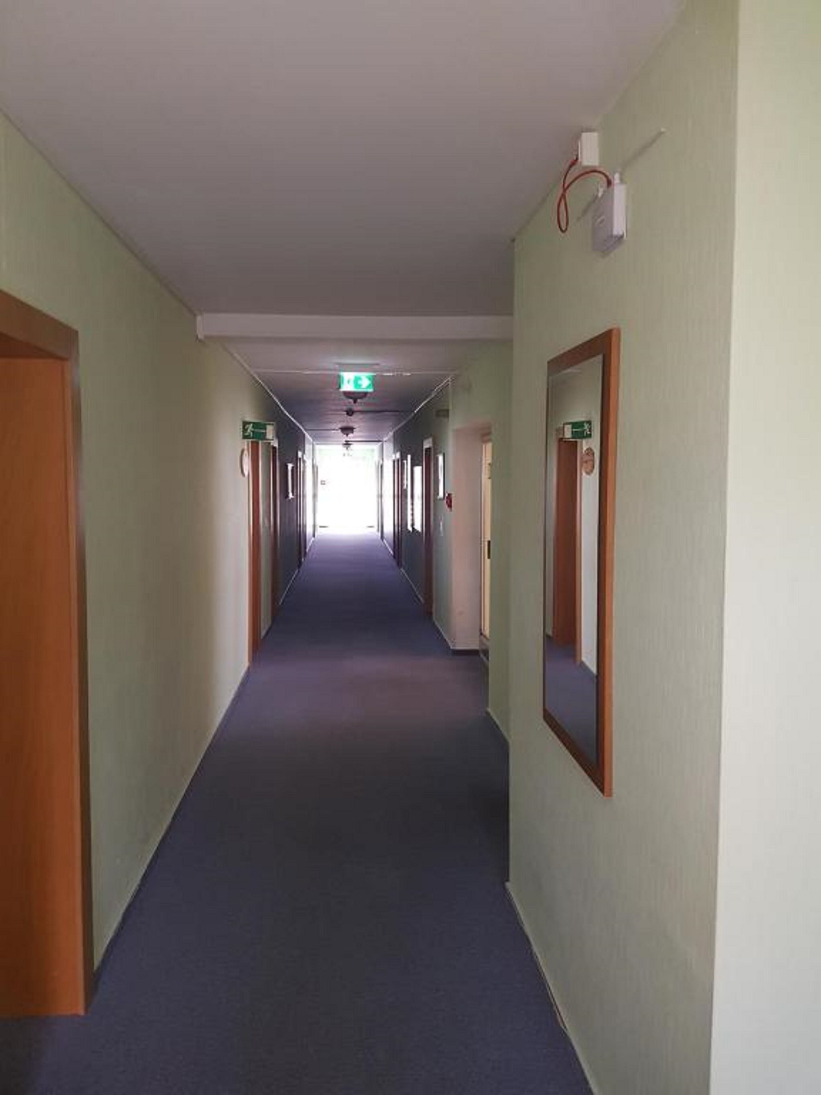 Un largo pasillo de hotel, estrecho, con puertas a ambos lados, suelo alfombrado y letreros de salida de emergencia.
