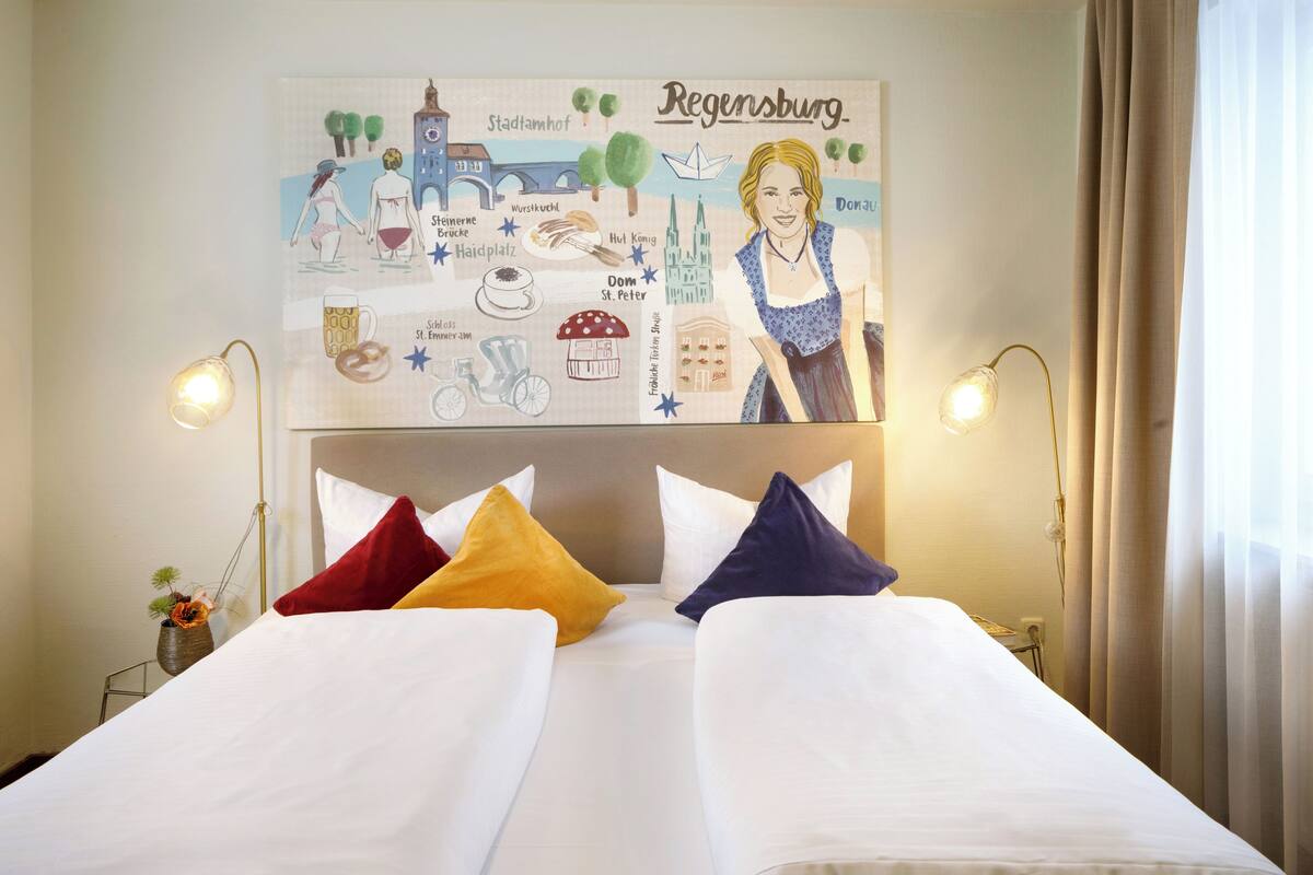 A hotel room with two beds, a large wall poster of Regensburg, and two bedside lamps.