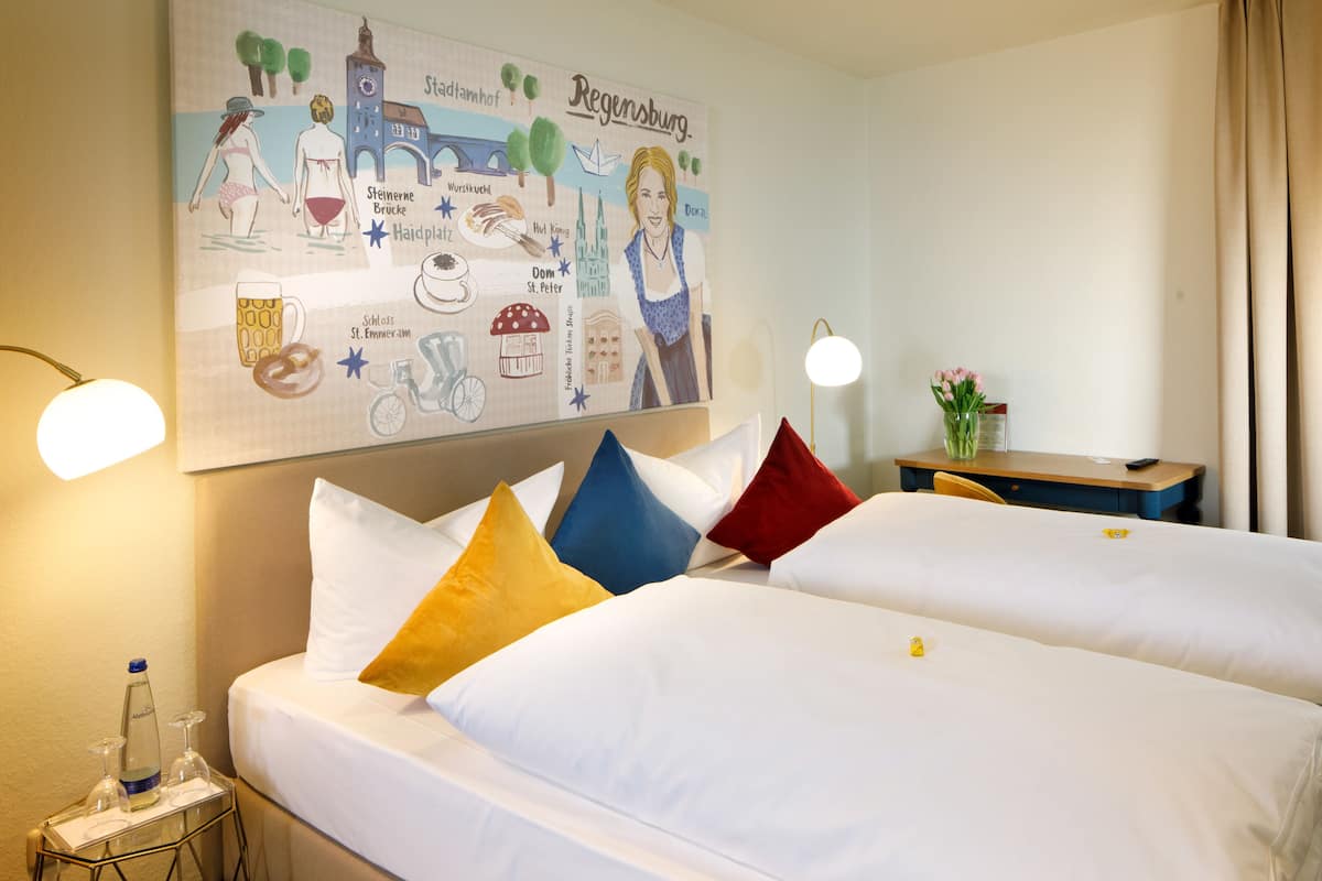 A hotel room with two beds, a nightstand with a bottle and glasses, a desk with a vase, and a wall with a cityscape illustration.