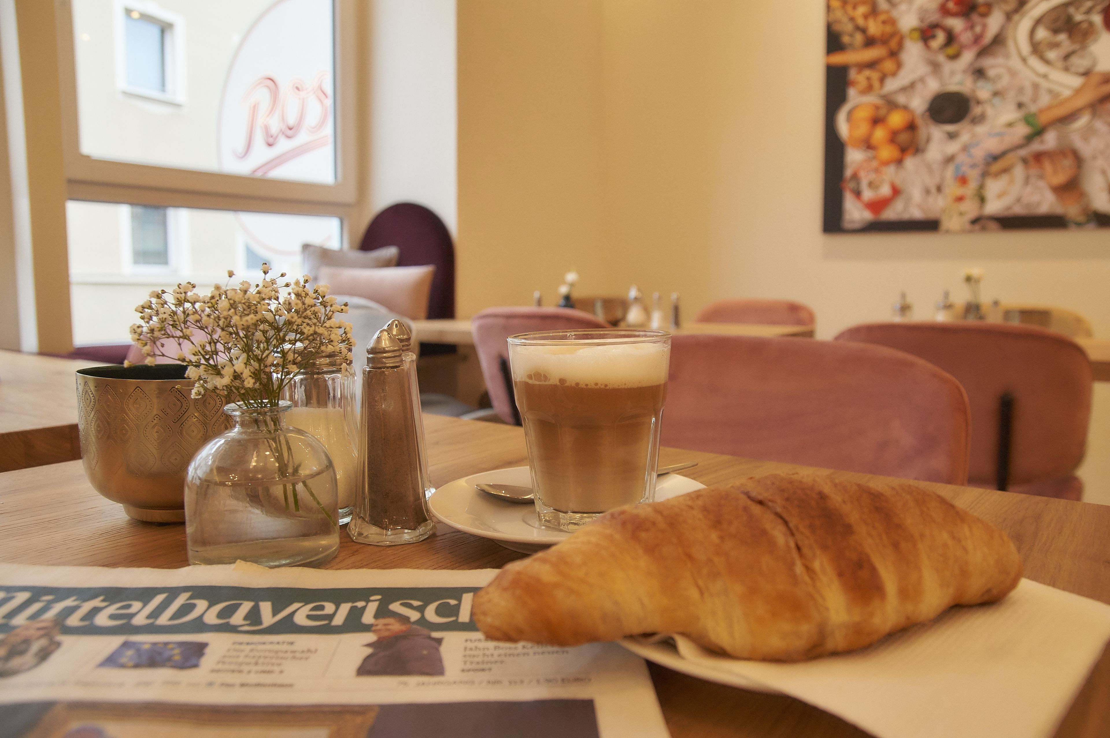 Una mesa con un croissant, una taza de café, un periódico y un florero con flores.