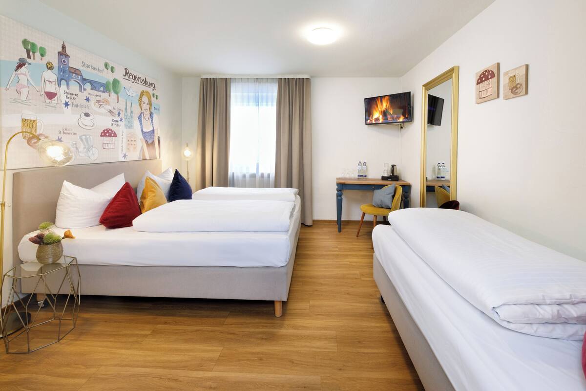 A hotel room with two beds, a desk, a chair, a TV, and a wall with a cityscape mural.