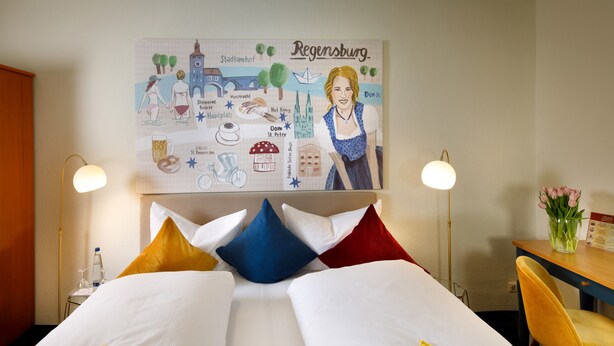 A hotel room with two beds, a large cityscape poster, two bedside lamps, a wooden nightstand, a chair, and a vase with flowers. - Rosi Boutique Hotel (Regensburg)