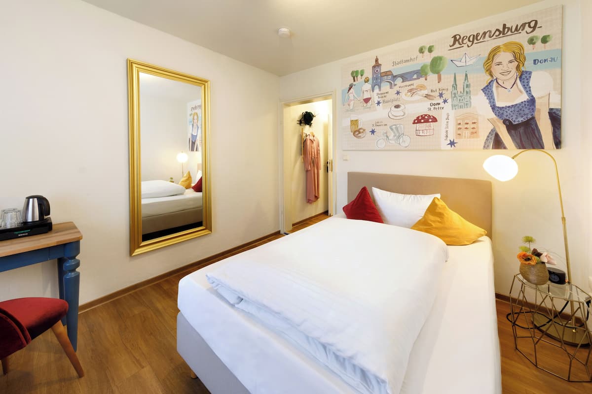 A hotel room with a bed, a desk with a coffee maker, a mirror, and a wall with a cityscape illustration.