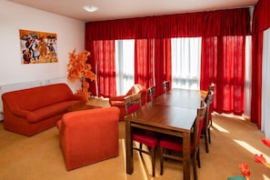A room with a red sofa, a wooden dining table with chairs, and a large window with red curtains.