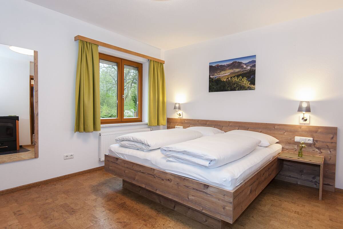 A bedroom with a wooden bed, a window with curtains, a nightstand with a lamp, and a wall-mounted picture.