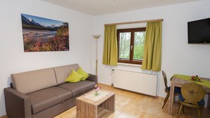 A modern living room with a sofa, a coffee table, a dining table with chairs, a window with curtains, a floor lamp, and a flat-screen TV. - Pension Leuprecht (Lechaschau)