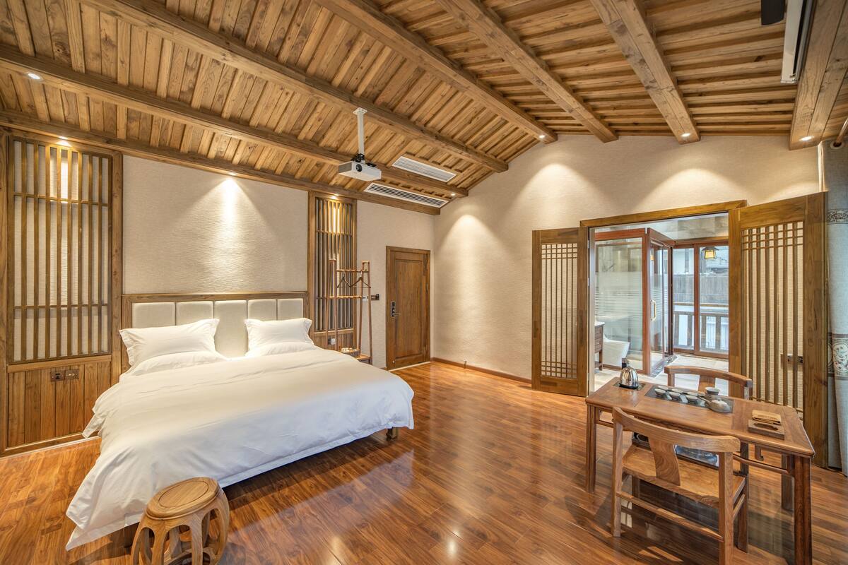 A spacious bedroom with a large bed, wooden ceiling beams, a wooden desk, and a traditional sliding door.