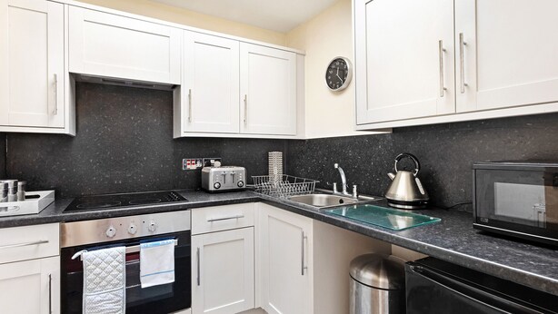 A modern kitchen with white cabinets, a black countertop, a stainless steel sink, a microwave, a kettle, and a toaster. - Granville Apartments (Harrogate)