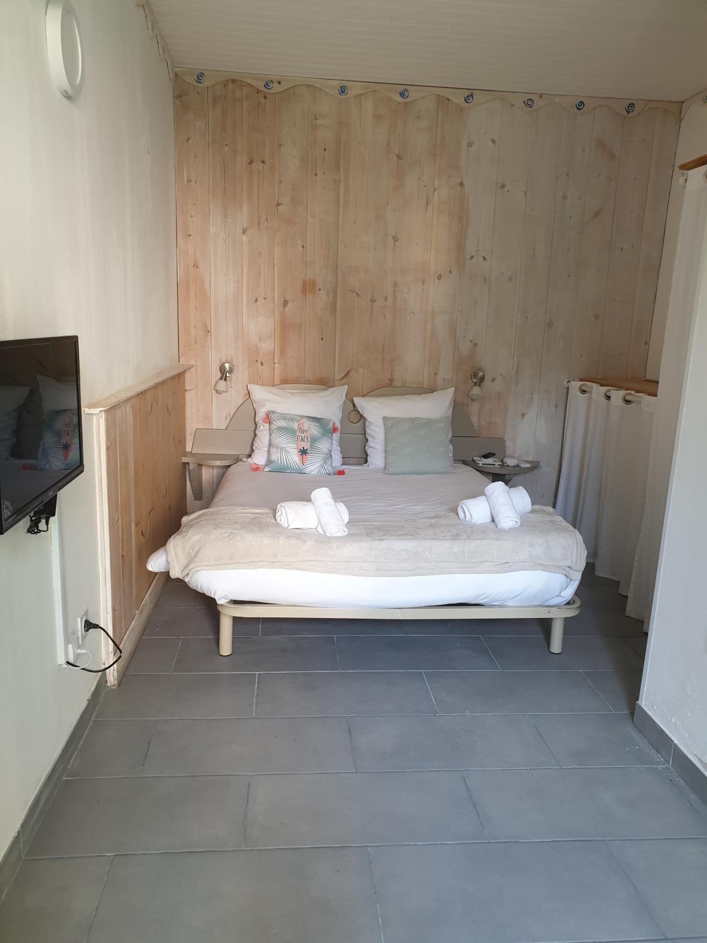 Een compacte, netjes ingerichte slaapkamer met een houten hoofdbord, een bed met witte lakens en handdoeken, een nachtkastje en een aan de muur bevestigde televisie.