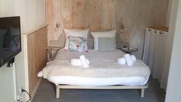 Een compacte, netjes ingerichte slaapkamer met een houten hoofdbord, een bed met witte lakens en handdoeken, een nachtkastje en een aan de muur bevestigde televisie.