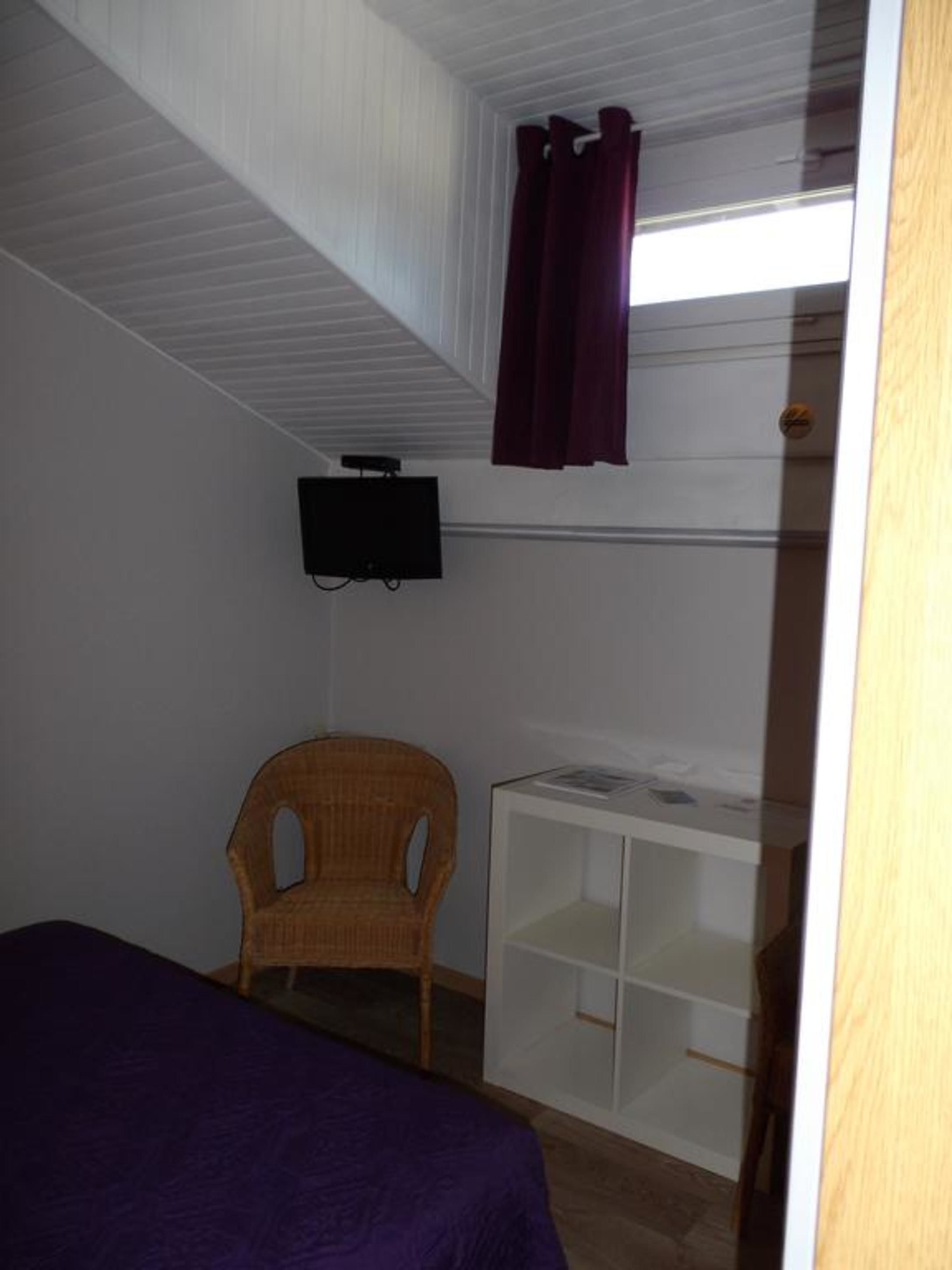 Une pièce comprenant un lit, une chaise en osier, une étagère blanche et un téléviseur fixé au mur.
