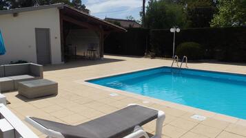 Une piscine avec une terrasse carrelée, un fauteuil de relaxation et un petit espace couvert avec des chaises.