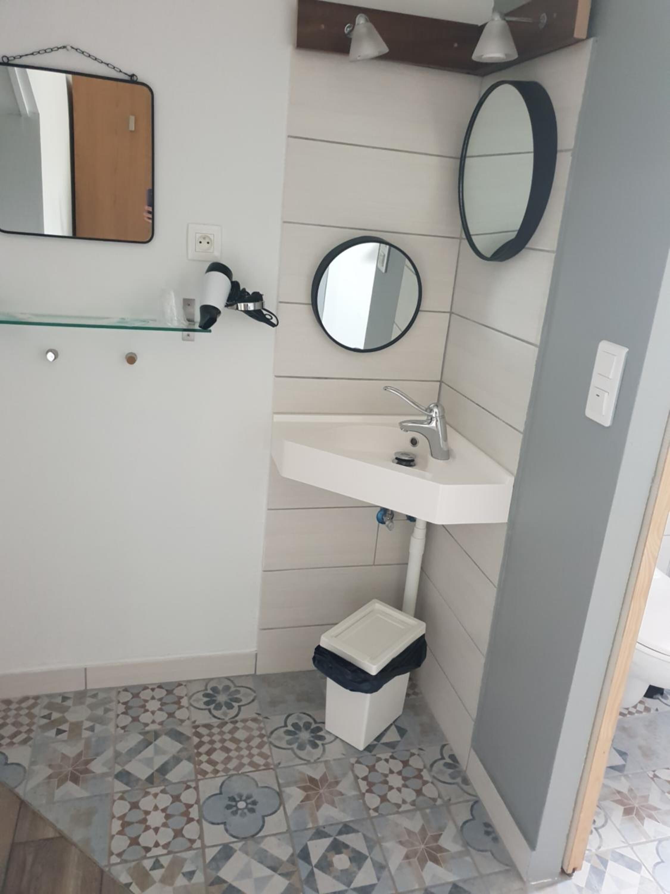 Casa de banho com lavatório branco, duas espelhas, prateleira de vidro e pavimento em azulejos estampados.