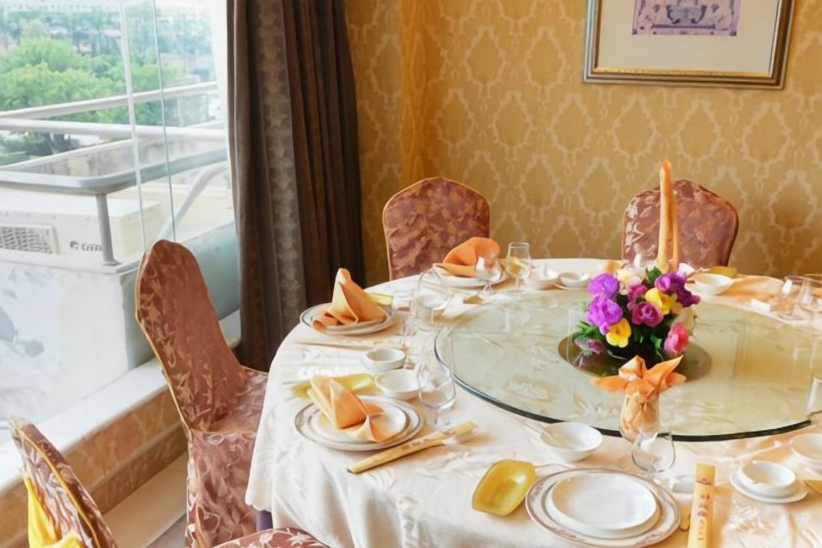 A dining table set for four with a floral centerpiece, a glass top, and a view of greenery outside.