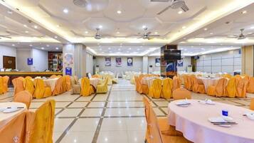 Un salón de banquetes con filas de mesas y sillas, ventiladores de techo y un escenario.