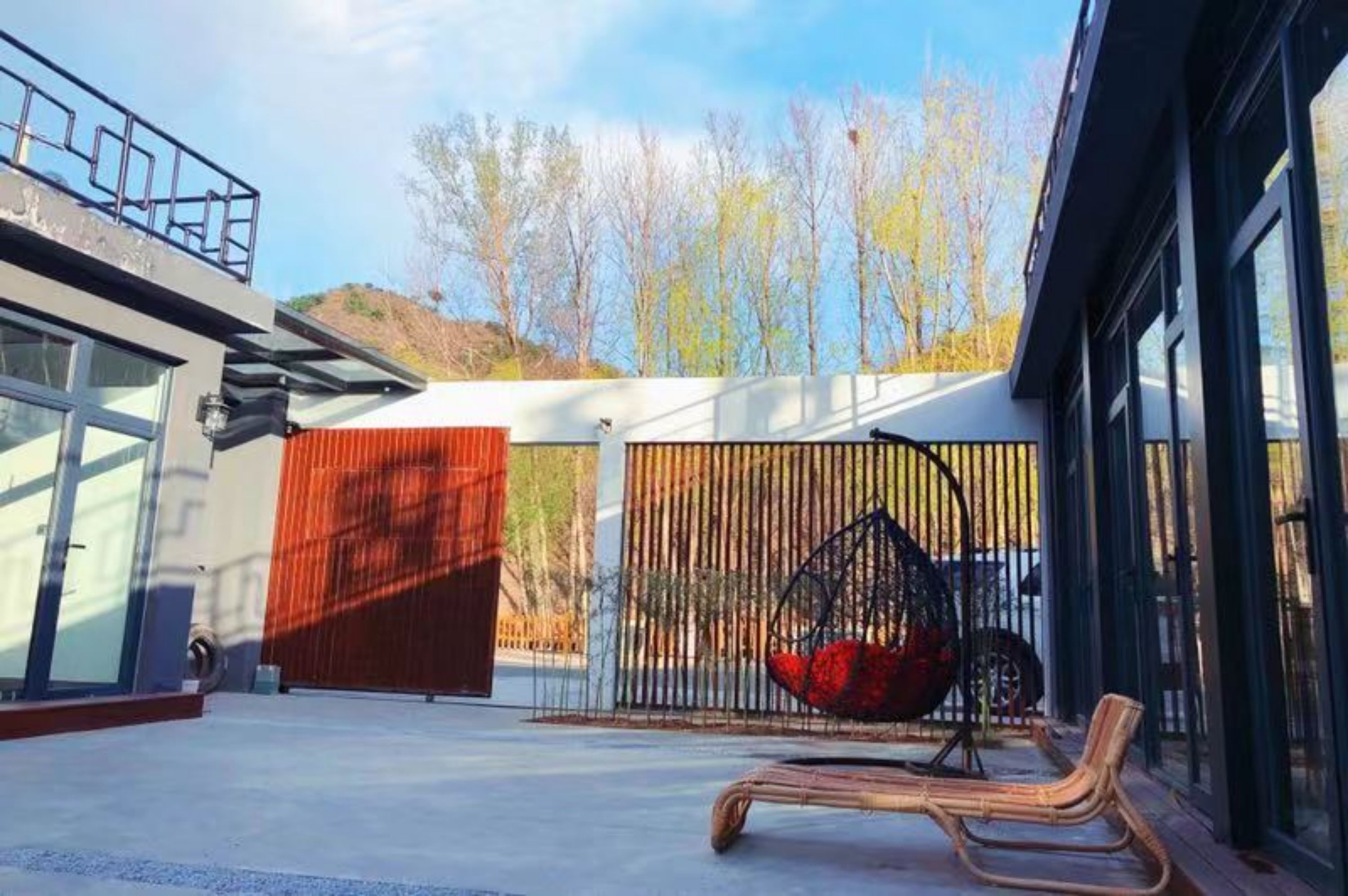 A modern outdoor seating area with a swing chair and a cushioned bench, enclosed by a metal fence and a red door.