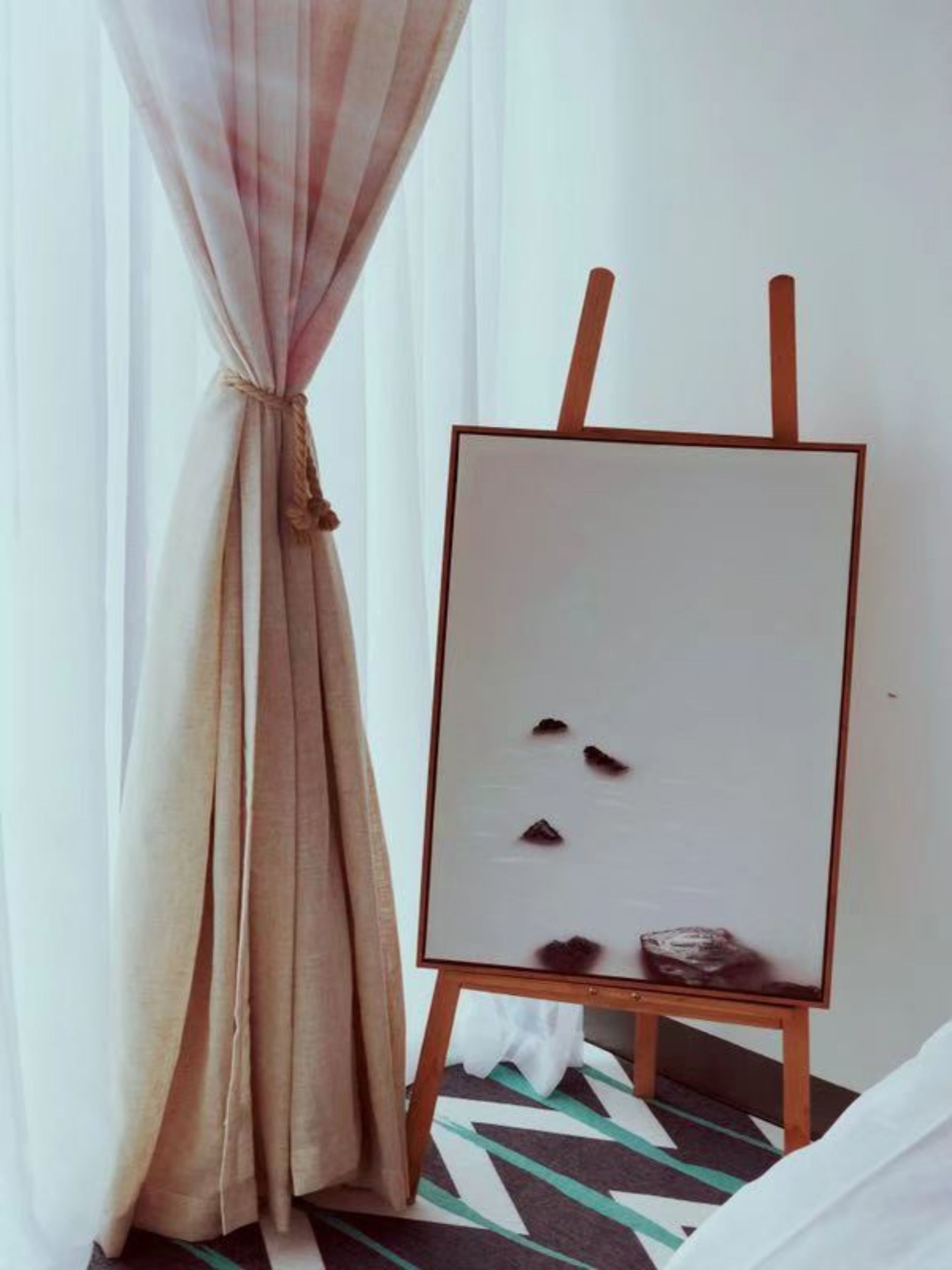 Una habitación con un caballete de madera apoyado en una pared, un tapete estampado y una cortina atada en la parte superior.
