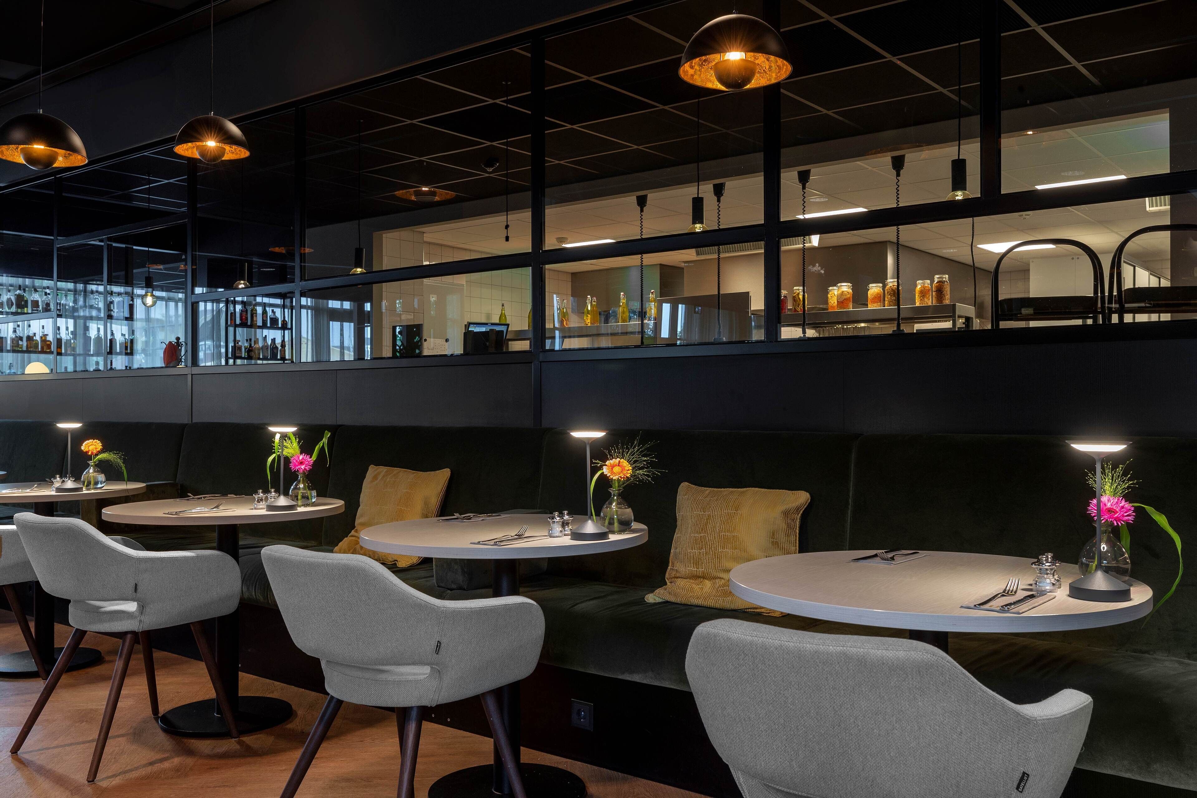 A modern restaurant with round tables and grey chairs, featuring a dark interior and visible kitchen area in the background.