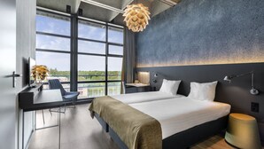 Una habitación de hotel moderna con una cama grande, un escritorio con una silla, una lámpara de escritorio y vistas al exterior a través de ventanas amplias.