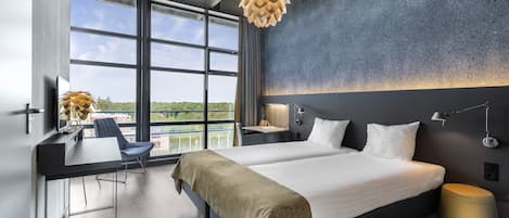 Una habitación de hotel moderna con una cama grande, un escritorio con una silla, una lámpara de escritorio y vistas al exterior a través de ventanas amplias.