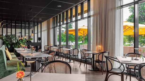 Un comedor moderno con mesas y sillas, ventanas amplias e interiores con plantas.
