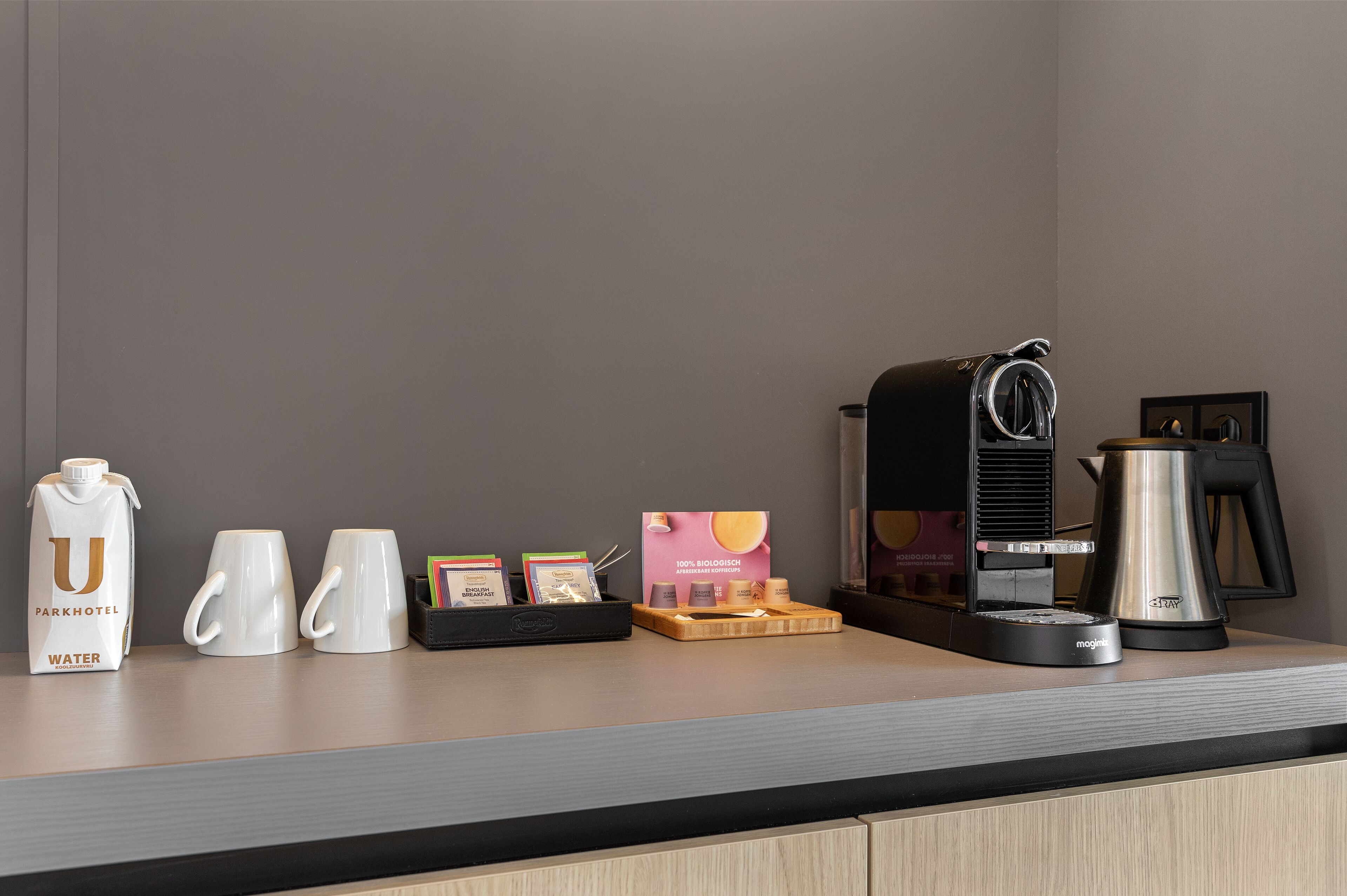 A coffee station with a water jug, two mugs, a tray with coffee pods, a coffee maker, and a kettle.