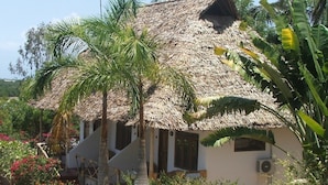 Ein Haus mit Strohdach und weißer Fassade, umgeben von Palmen und blühenden Pflanzen.
