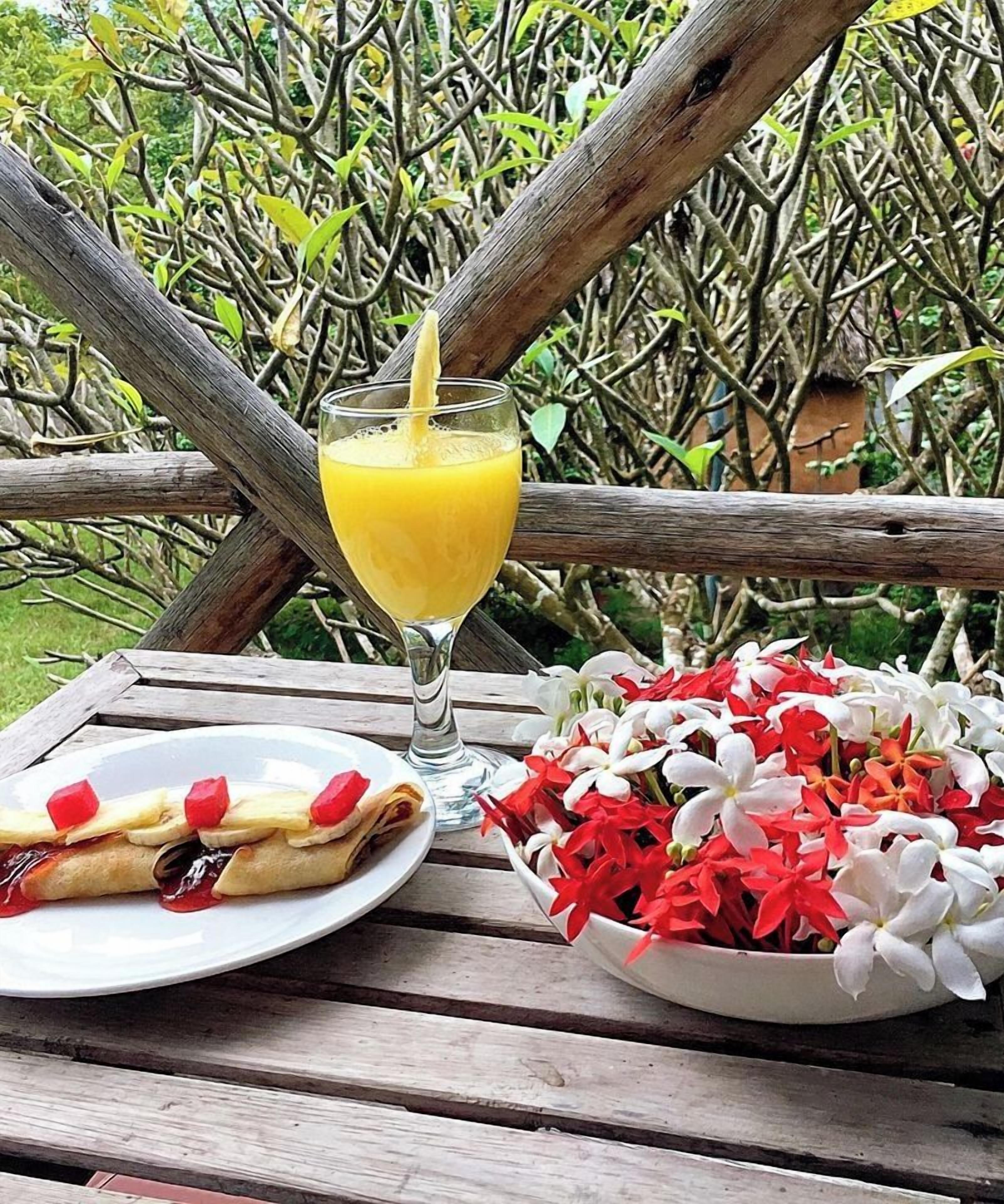 Una mesa de madera con un vaso de jugo de naranja, un plato de comida y un cuenco de flores.