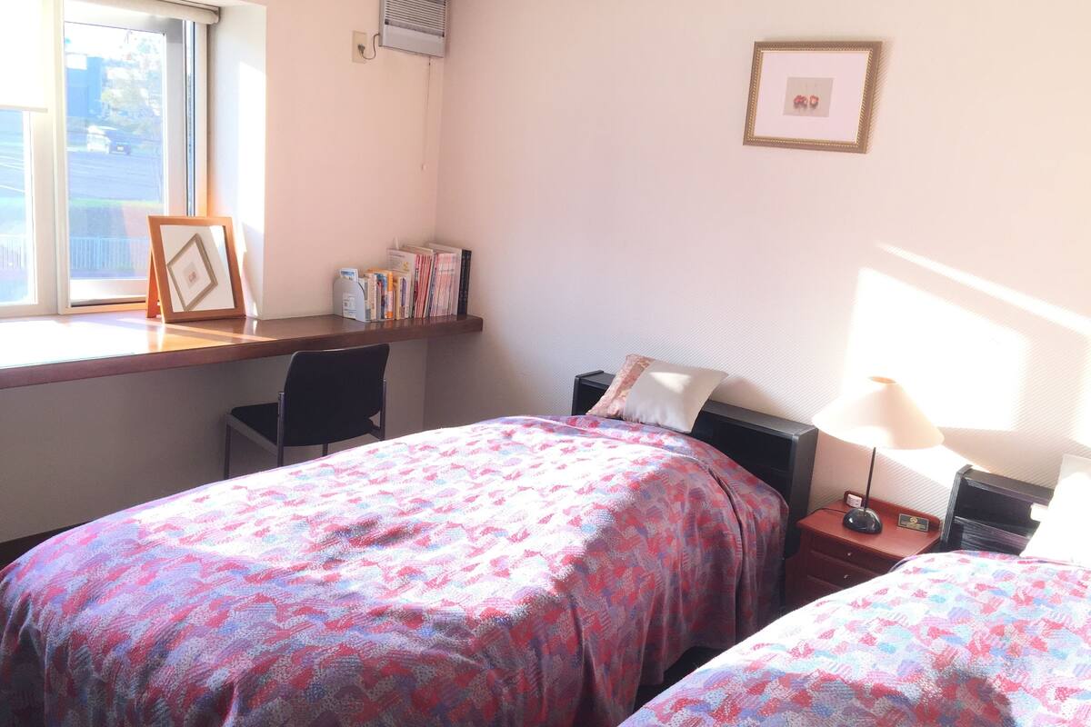 A bedroom with two beds, a desk, a chair, a nightstand, and a window.