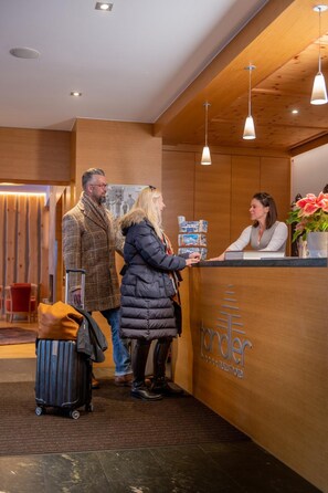 A reception desk with a wooden finish and a logo, a person standing at the desk, and a luggage trolley in the foreground.