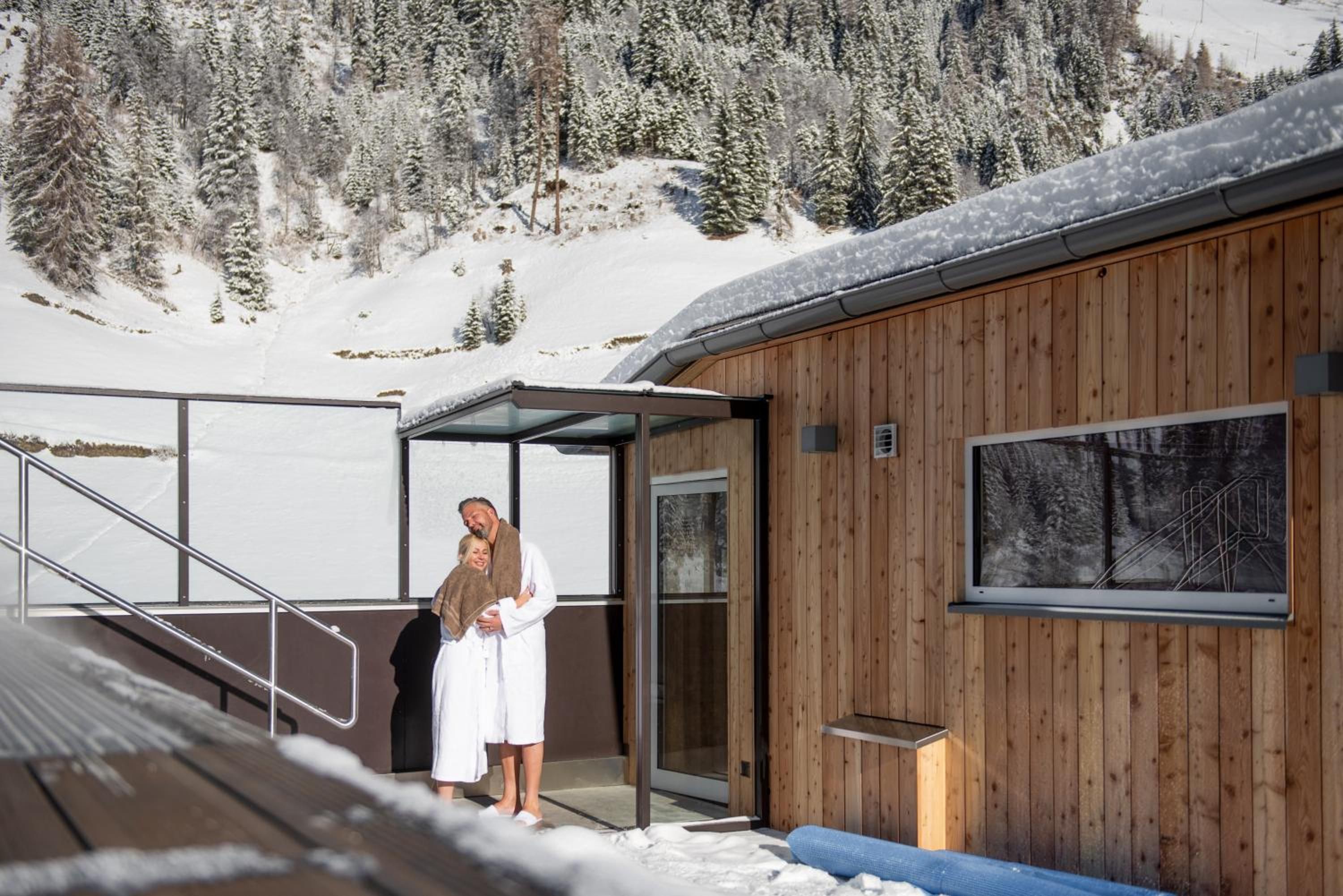 A wooden cabin with a glass door, a metal staircase, and a view of snow-covered mountains.