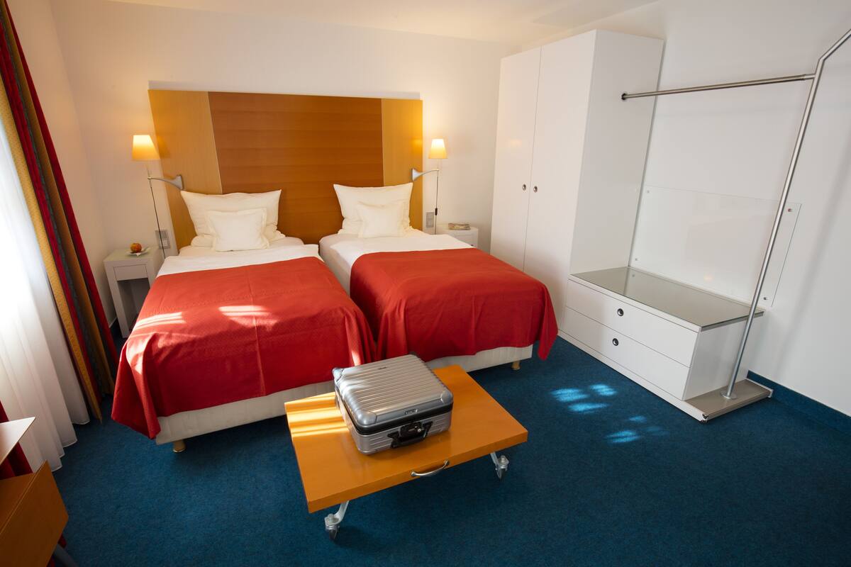 Une chambre d’hôtel avec deux lits, une petite table sur laquelle est posée une valise, une armoire et une table de chevet avec une lampe.