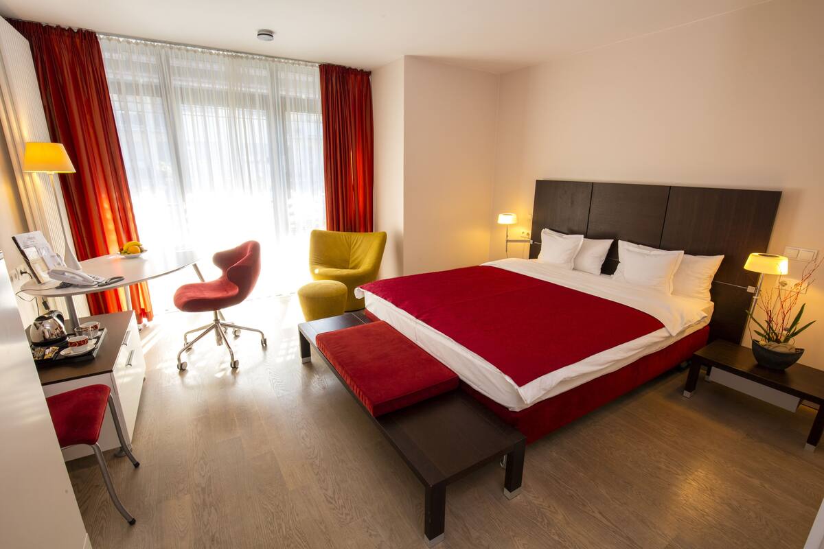 Une chambre d’hôtel avec un grand lit, un bureau avec une chaise, une petite table et une chaise rouge.