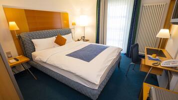 A hotel room with a large bed, two bedside tables, a desk with a chair, a telephone, and a radiator.