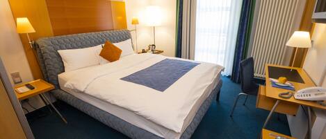 A hotel room with a large bed, two bedside tables, a desk with a chair, a telephone, and a radiator.