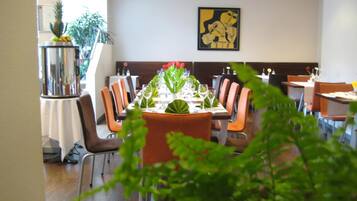Ein Restaurant mit einem langen Tisch, gedeckt für ein Essen, umgeben von Stühlen und einer Pflanze im Vordergrund.
