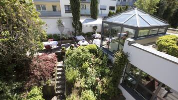 A rooftop garden with a glass greenhouse, outdoor seating, and surrounding greenery.