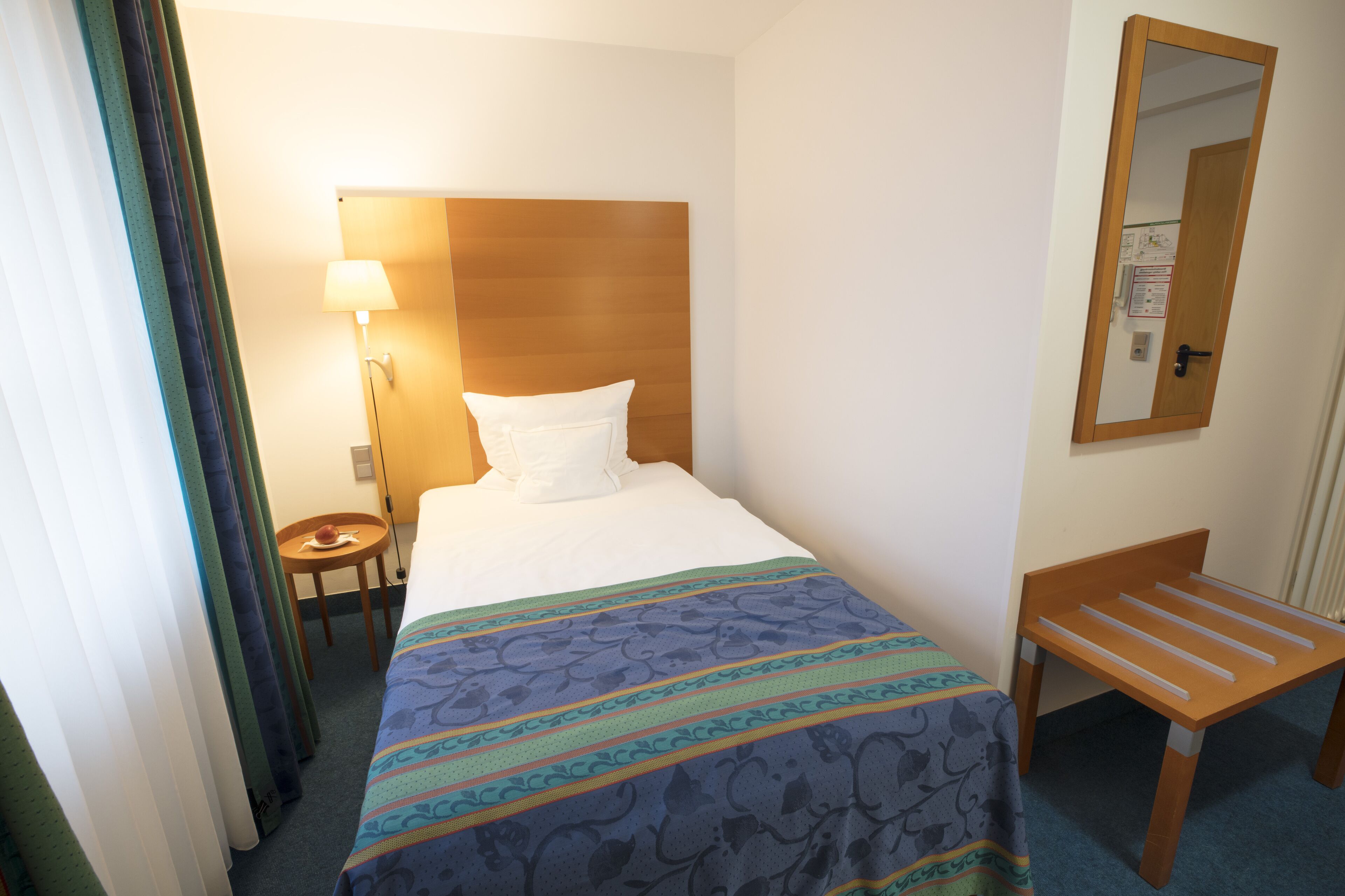 A hotel room with a bed, a bedside table with a lamp, a wooden bench, and a mirror on the wall.