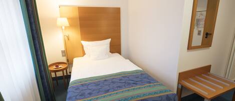 A hotel room with a bed, a bedside table with a lamp, a wooden bench, and a mirror on the wall.