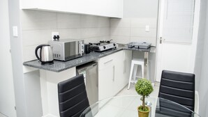 A compact kitchen with a coffee maker, toaster, and microwave on the countertop, a dishwasher, and a small dining area with two chairs and a table. - Cape Town Micro Apartments (Cape Town)
