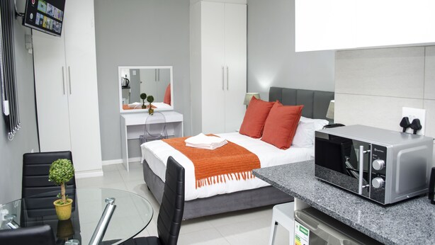 A modern bedroom with a bed, a desk, a chair, a microwave, and a TV. - Cape Town Micro Apartments (Cape Town)