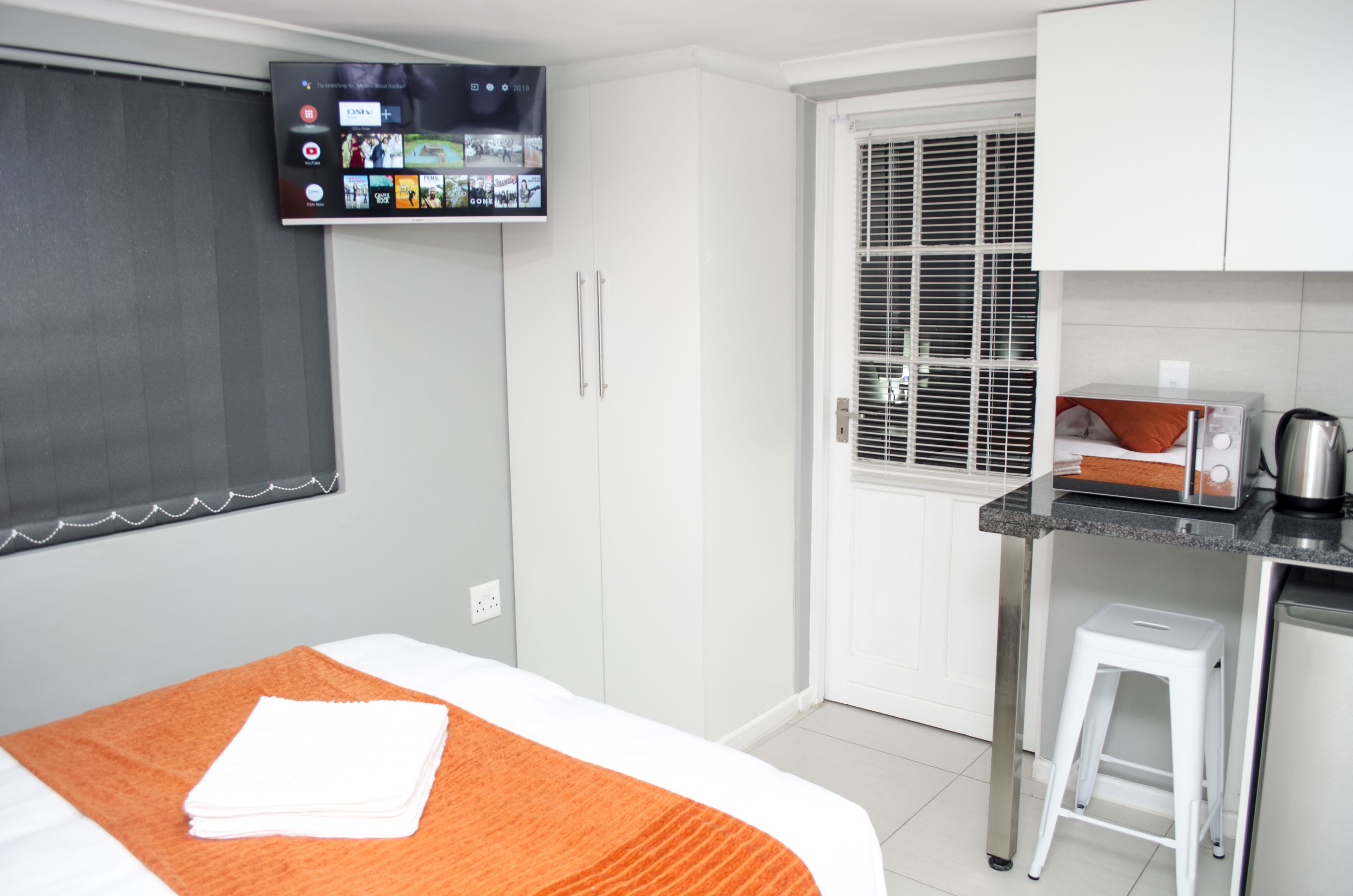 A hotel room with a bed, a TV mounted on the wall, a kitchenette, and a window with blinds.