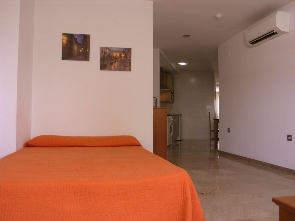 Una habitación con una cama, dos cuadros en la pared, un equipo de aire acondicionado y una zona de cocina al fondo.